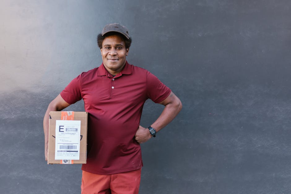 A male flower delivery courier from Cowleyflowers.co.uk stands outdoors against a plain dark grey wall, smiling at the camera while holding a small cardboard parcel with a delivery label. He is dresse