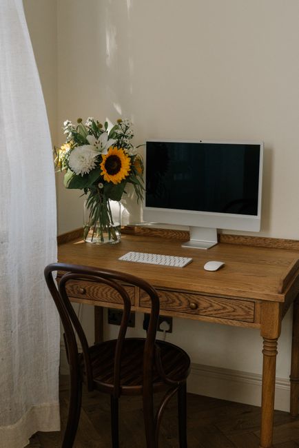A clear glass vase holding a fresh floral arrangement positioned on a wooden desk in a well-lit room. The bouquet features large white chrysanthemums, vibrant yellow sunflowers, and smaller white and 