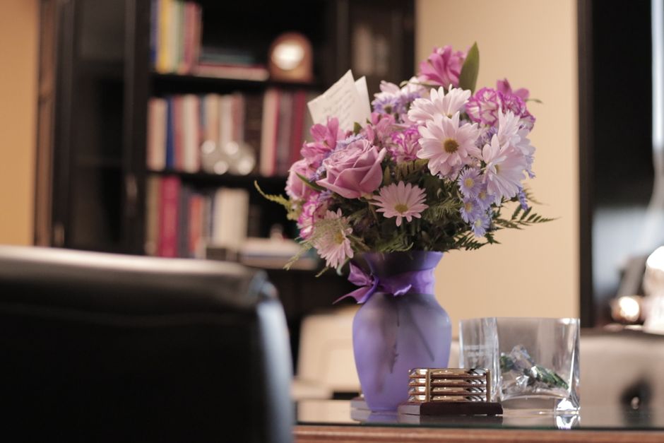 A vibrant bouquet of fresh flowers arranged in a purple ceramic vase, featuring pink roses, light purple daisies, and delicate purple and white filler blooms, with lush green fern leaves adding contra