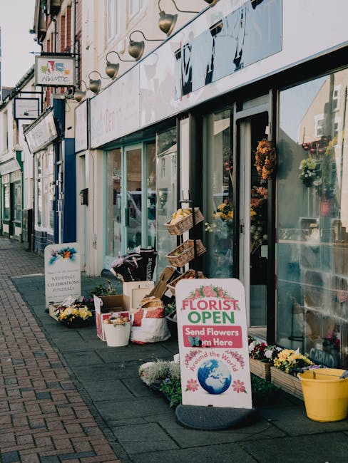 Exterior view of a flower shop on Cowley Road with large glass windows displaying floral arrangements inside. Outside, there are buckets and baskets filled with fresh, colorful flowers, including whit