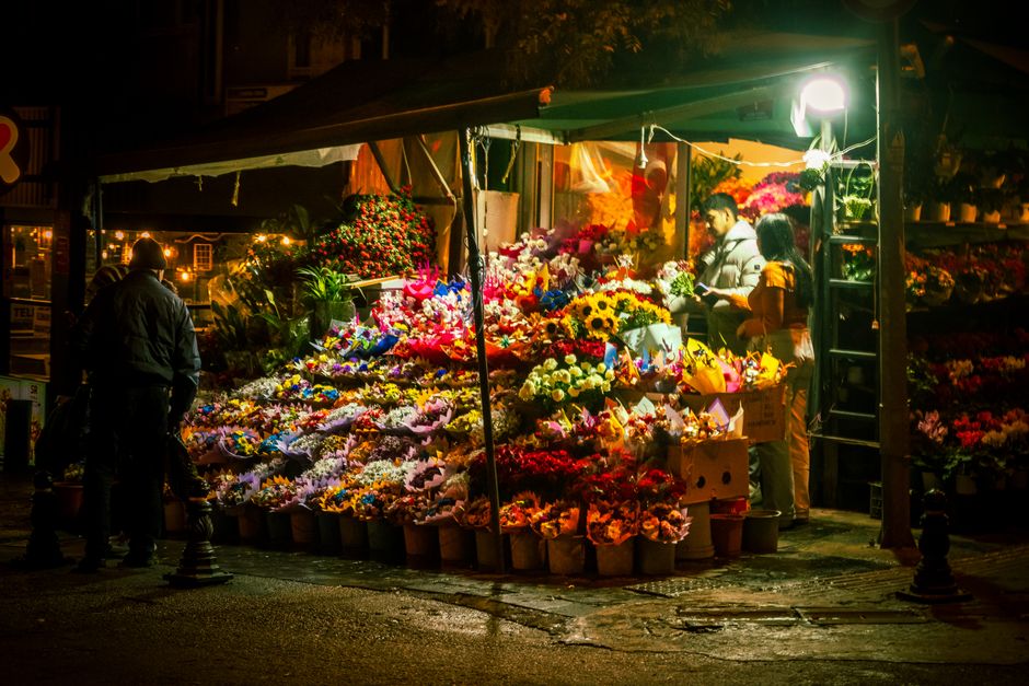 A flower stall at night illuminated by bright lighting, displaying a vibrant array of fresh flowers arranged in bouquets and containers. The flowers include sunflowers, roses, lilies, and various colo