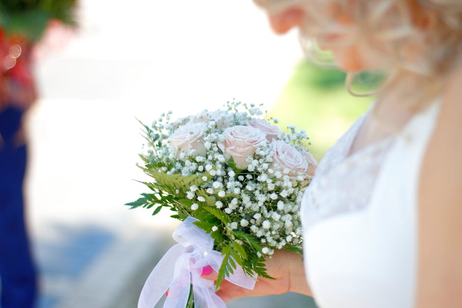 A close-up of a bride holding a bouquet arrangement featuring soft pink roses and delicate white baby's breath flowers, accented with green fern and foliage. The bouquet is wrapped with a white satin 