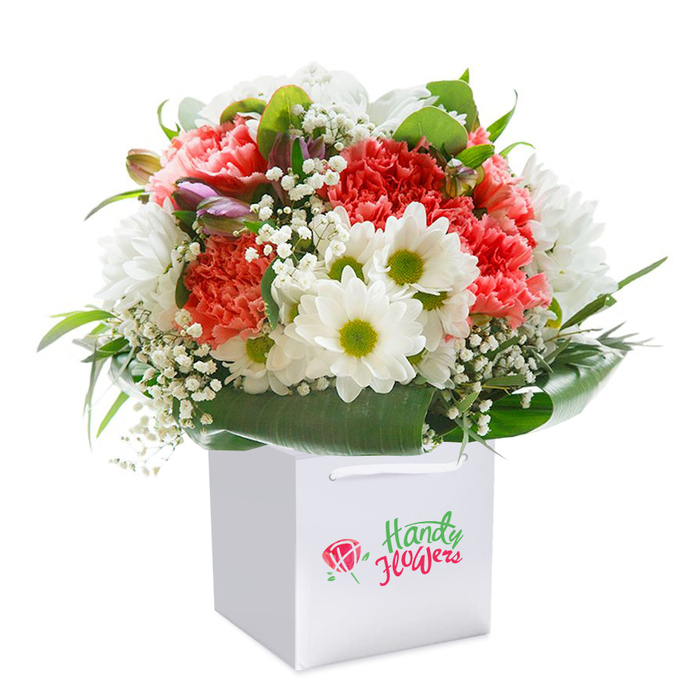 In the center of this luminous image, a beautifully balanced bouquet rests within a white, square gift bag-its composition a gentle salute to Cowley's tradition of vibrant, heartfelt floristry. Clusters of coral and peach carnations fan outward, their ruffled petals cascading with softness and generosity. Dotted among the main blossoms, generous sprays of bright white daisy chrysanthemums, each sporting a lime-yellow center, infuse the arrangement with a freshness that feels at home along Cowley Road's cheerful storefronts. Baby's breath is liberally interlaced, creating a floaty, airy impression, while hints of purple-tinged buds at the bouquet's edge provide added intrigue and depth-much like Cowley's lively mix of old and new. The base of the bouquet is gently held by a collar of broad, deep green leaves, their glossy finish nodding to the leafy pockets and gardens peppered throughout the area. The white gift bag, practical yet elegant with its twin ribbon handles, makes this bouquet ideal for collecting in person or sending straight to a loved one's door. Expertly arranged and photographed under clear, even light, the bouquet showcases both a vibrant sense of plenty and the personal care that defines Cowley Flowers. It's the perfect gift for marking joyful milestones-delivered fresh and full of heart throughout Cowley.