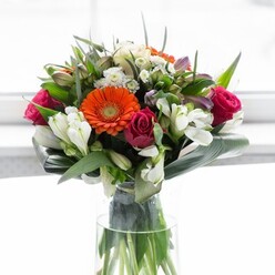 Expertly composed by Cowley's floral artisans, this bouquet is a celebration of colour, fragrance, and nature's joyful abundance. Creamy peach gerberas and glowing orange varieties sit gracefully together, their soft petals resting against the playful depth of magenta roses. Interwoven throughout are pure white alstroemeria and wisps of delicate filler blossoms, adding a gentle lightness that recalls early sunlight filtering through the trees of Cowley Marsh Park. Encircling the arrangement, broad, glossy aspidistra leaves form a protective collar, their dark emerald tones grounding the vibrant hues above and echoing the neighbourhood's natural charm. Splashes of lighter pink and additional fresh greenery create depth and movement, encouraging the eye to wander through the bouquet's intricate tapestry. The tall glass vase reveals strong, healthy stems, while a sunlit, minimalist background ensures every flower shines. Perfect for a spring celebration, welcoming a new neighbour to Oxford, or brightening a day in Iffley, this arrangement is a testament to Cowley Flowers' commitment to quality and community spirit, marrying tradition with modern design.
